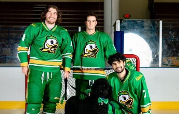 picture of henry andres dressed in full Oregon Ducks hockey uniform standing on a hockey ice, next two two other players and a black dog.
