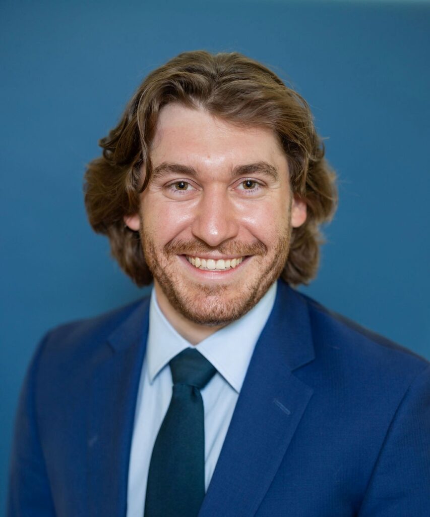 professional headshot of henry andres wearing a suit coat and tie sitting in front of a blue background.